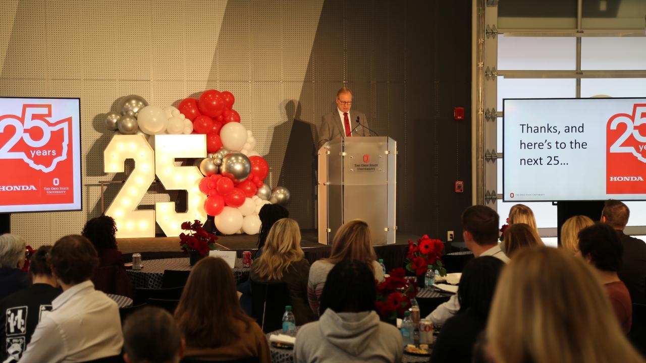 A speaker stands at a podium during an event celebrating 25 years of partnership between Honda and The Ohio State University, with large “25” decorations and red, white, and silver balloons on stage.