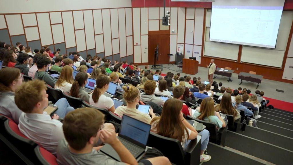 Students seated in Schoenbaum Hall listening to a professor presenting slides on a projector screen. Many students have laptops open and are taking notes.