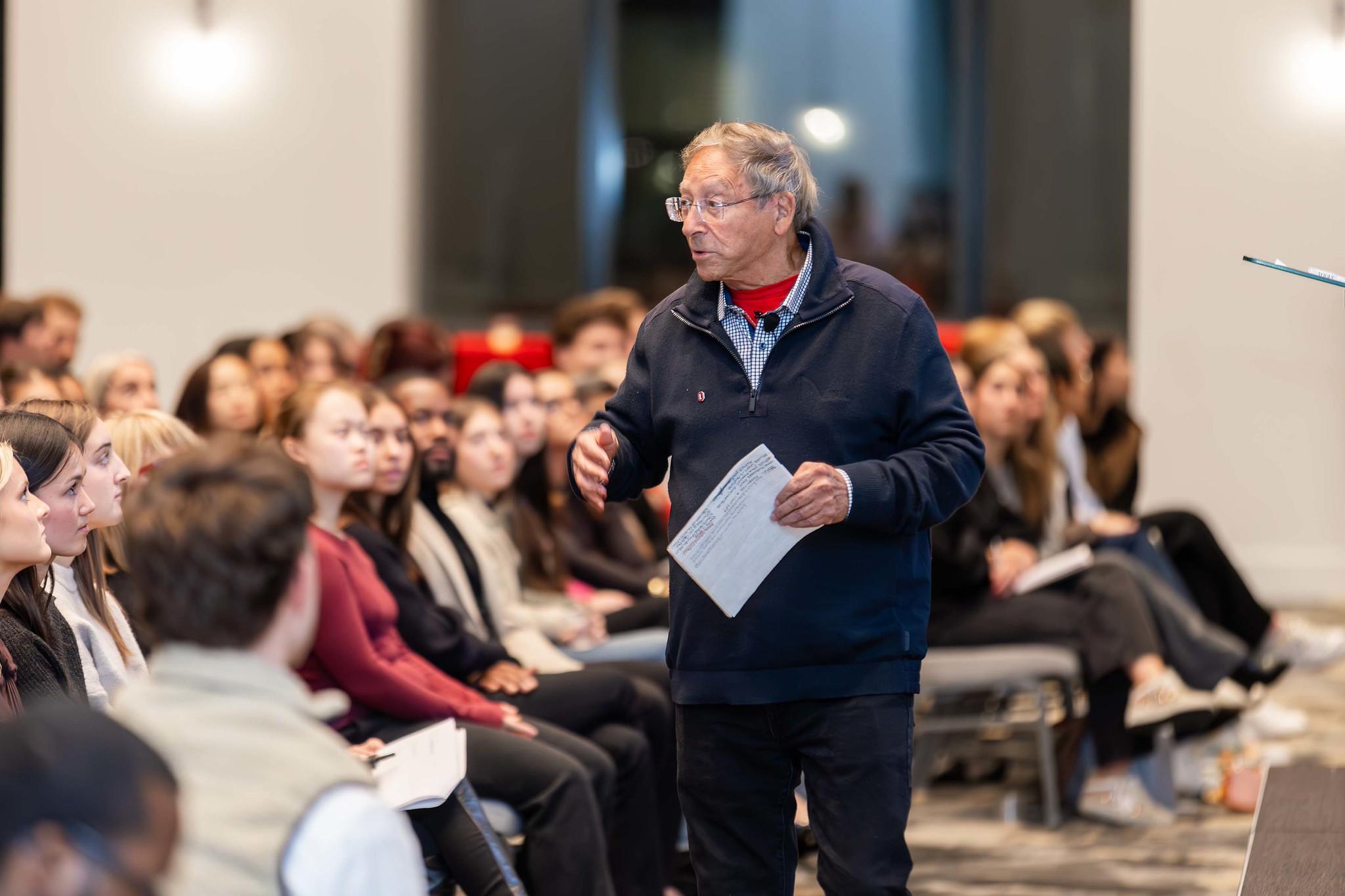 Stuart Weitzman standing and speaking to a seated audience in a conference or seminar setting, holding a sheet of paper.