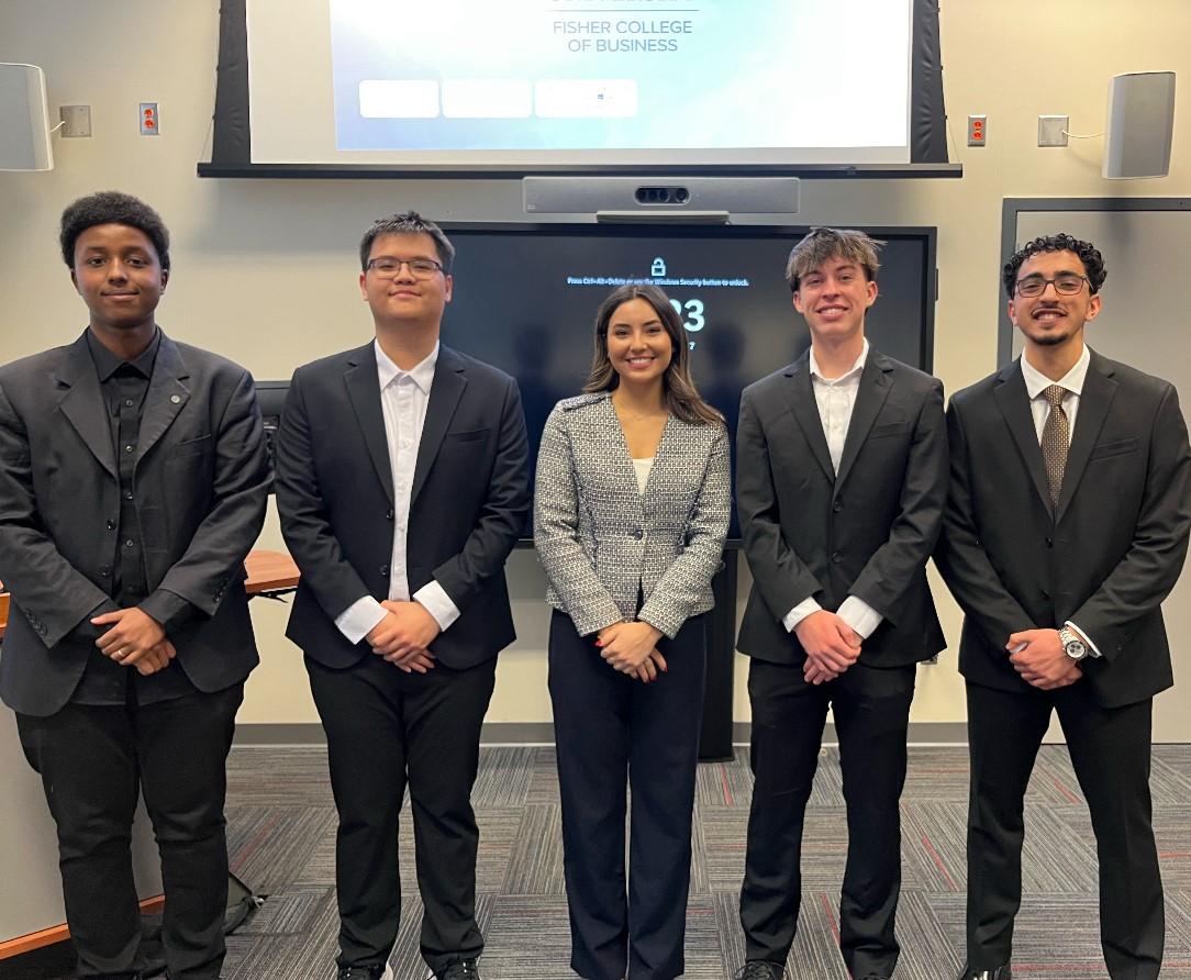 group of five students in business suits in front of screen in classroom