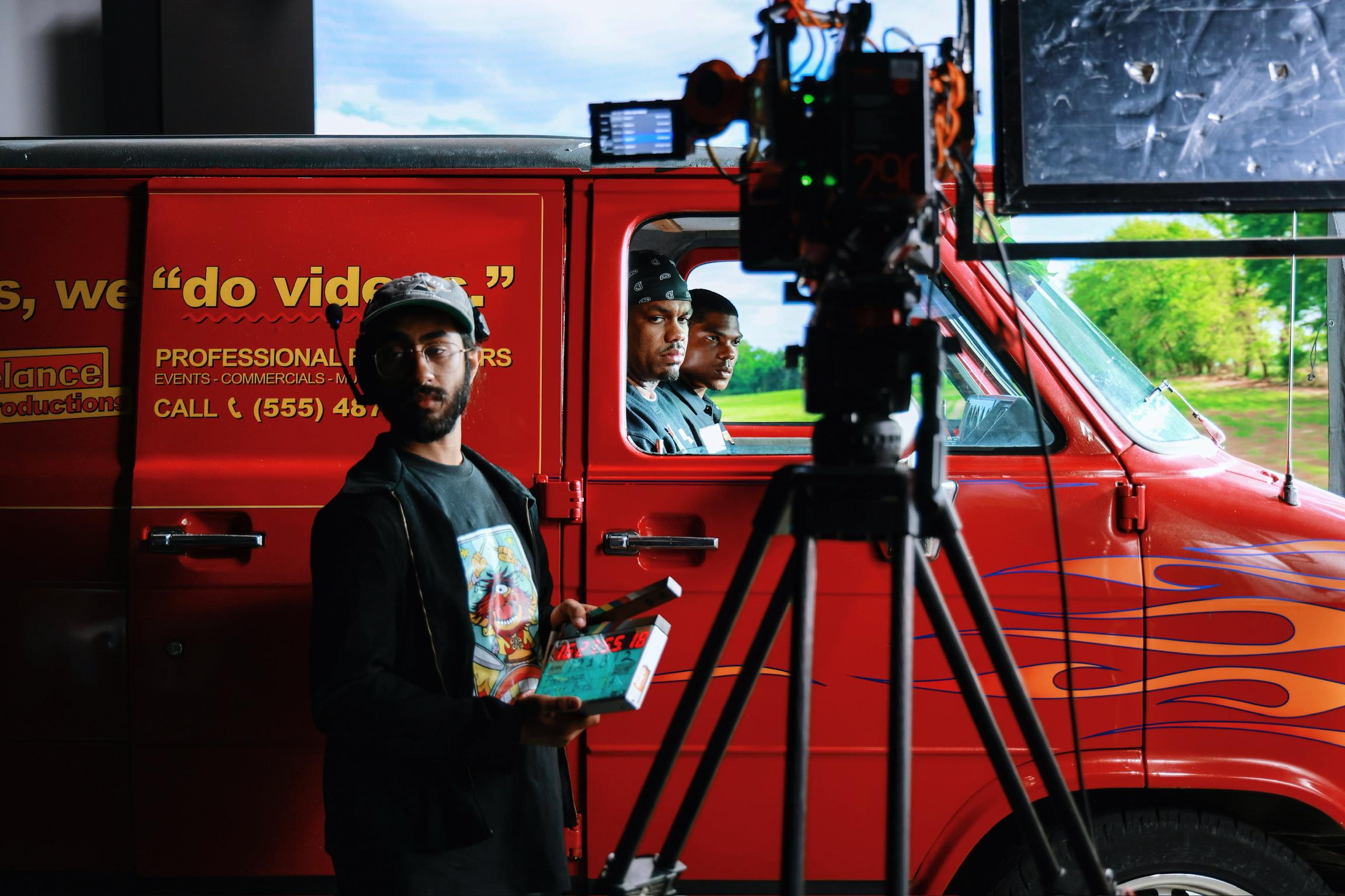 A cameraman stands beside a movie camera in front of a van with two actors inside on the set of a TV pilot.