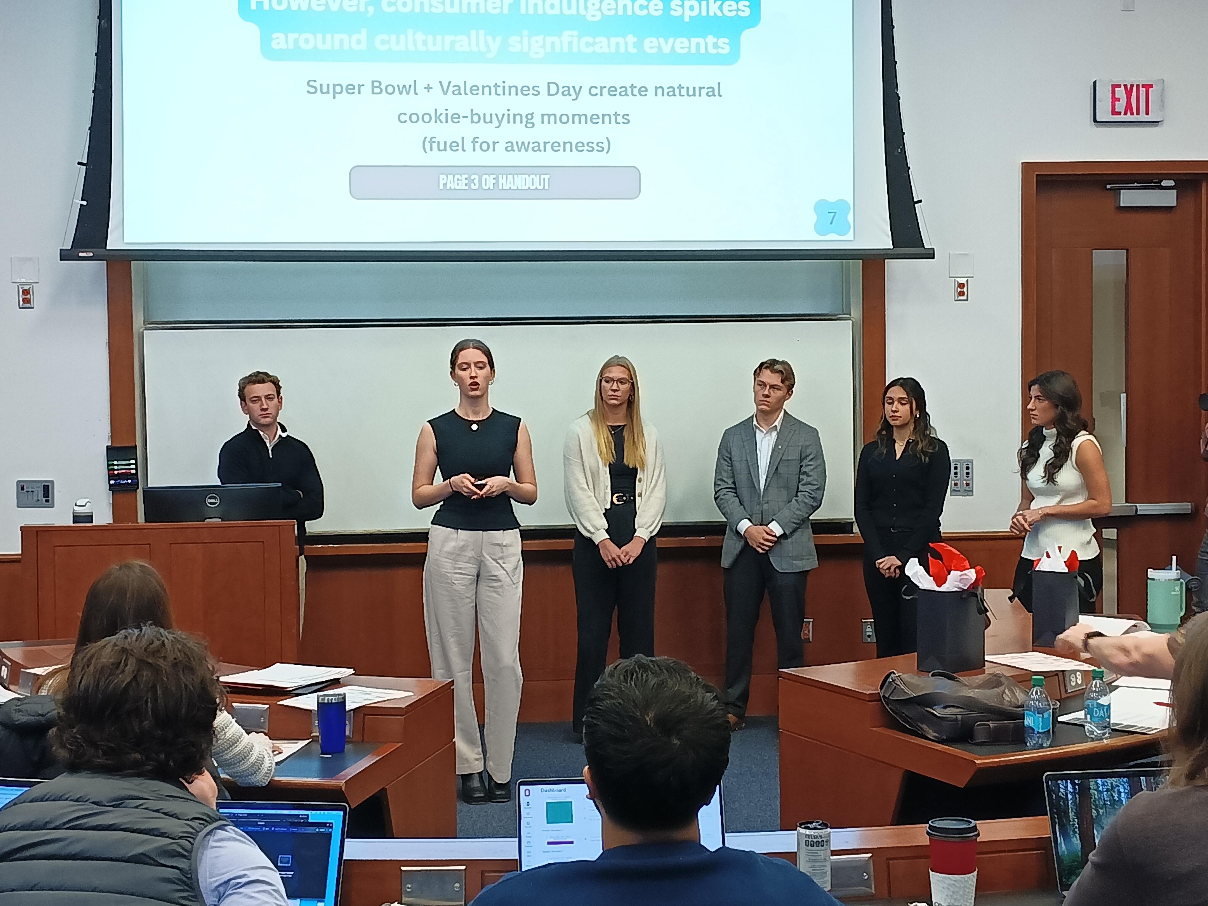 Group of students in front of classroom with presentation screen pulled down.