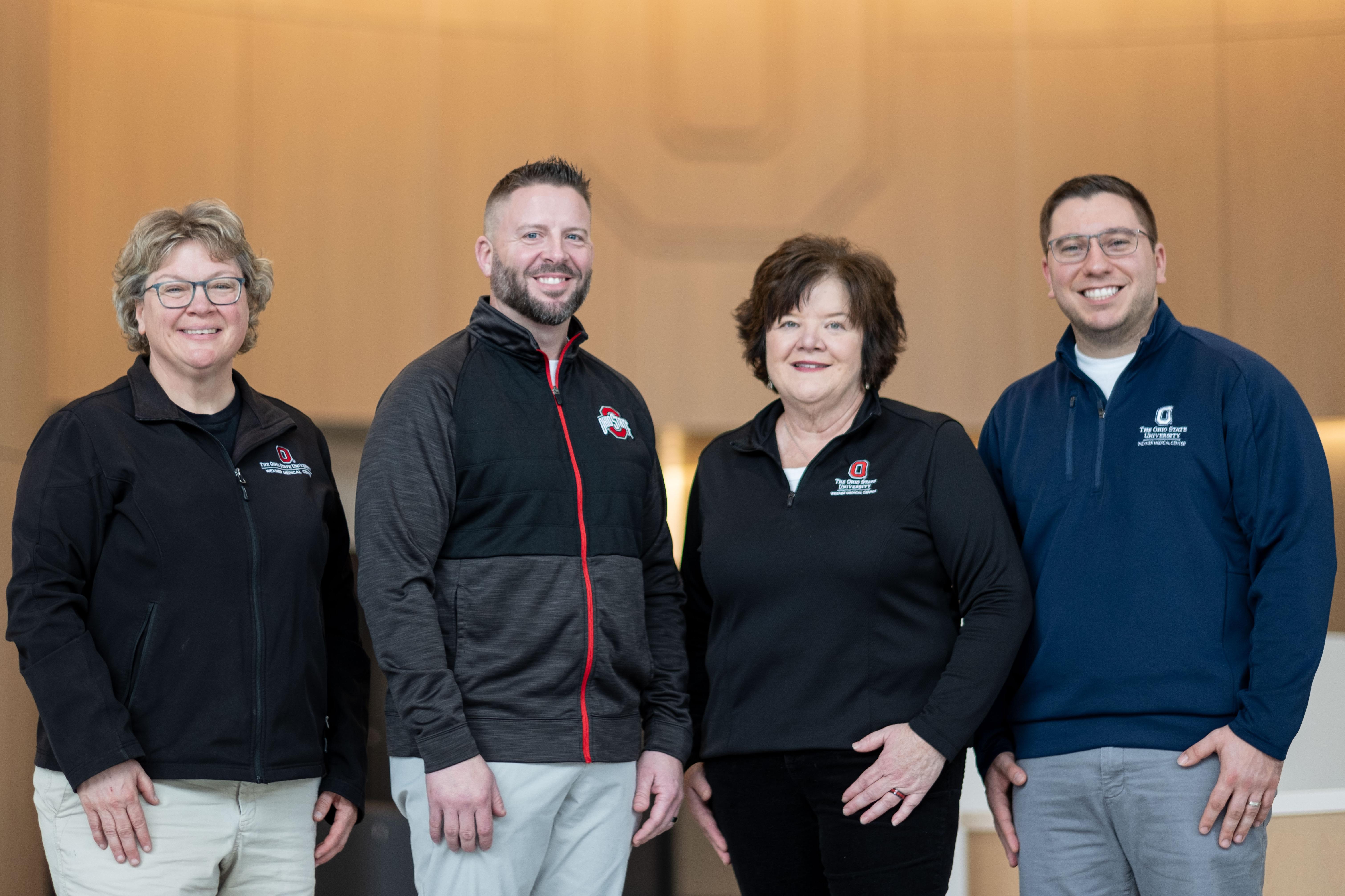 MBOE alumni from left to right, Lori Abshire, Calen Bowshier, Devanie Gossett and Mike Turnbull pose in the lobby of University Hospital in front of a wood paneled wall with a block O engraved in it.