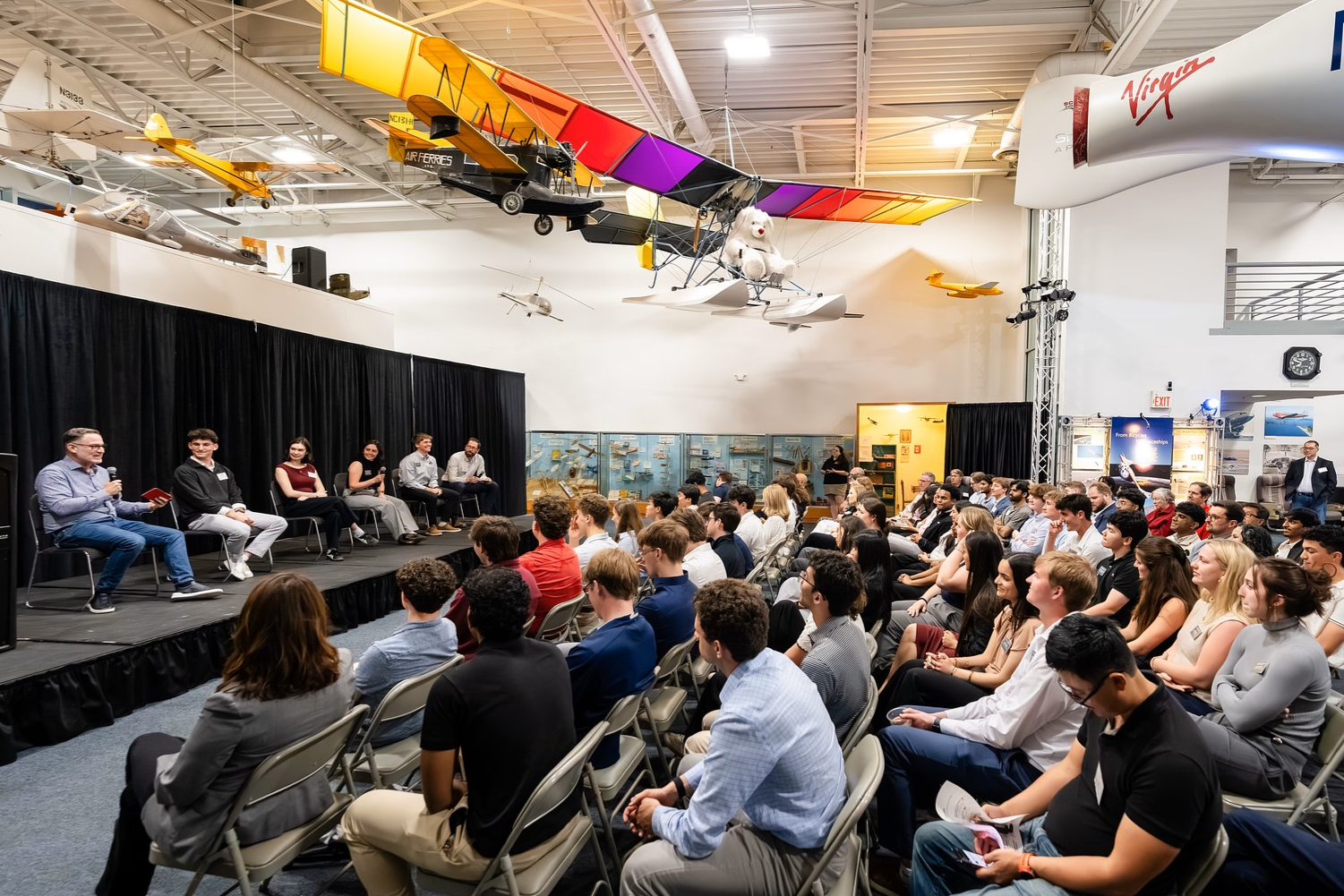 A group of alumni and students are seated on chairs in front of a panel of six people incise an aviation museum with replicas of airplanes overhead.