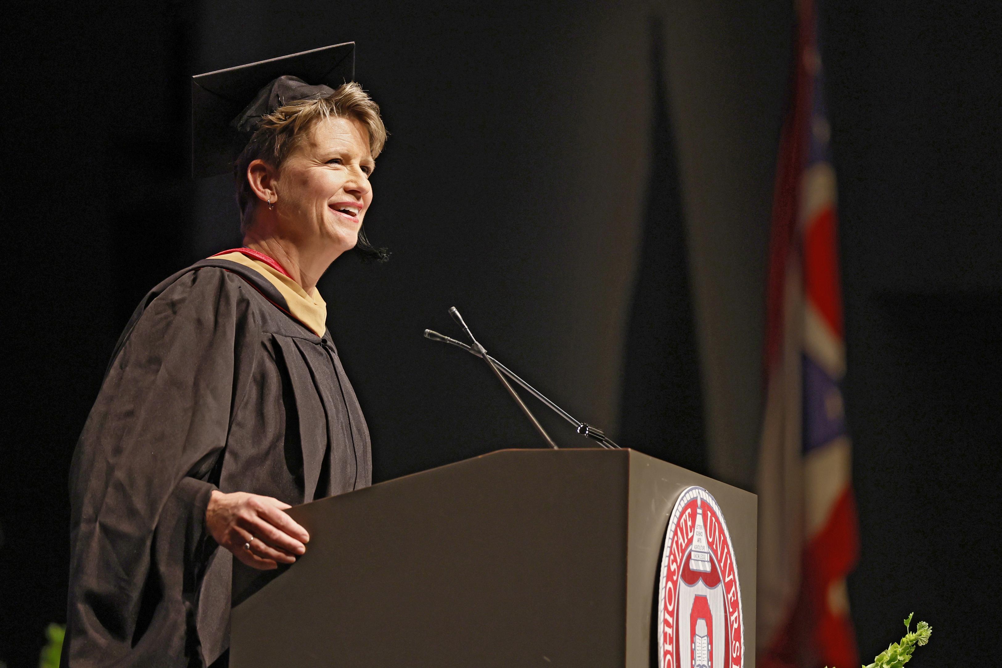 Melinda Whittington in a black cap and gown and speaking into a microphone at the podium during the Pre-Commencement Ceremony.