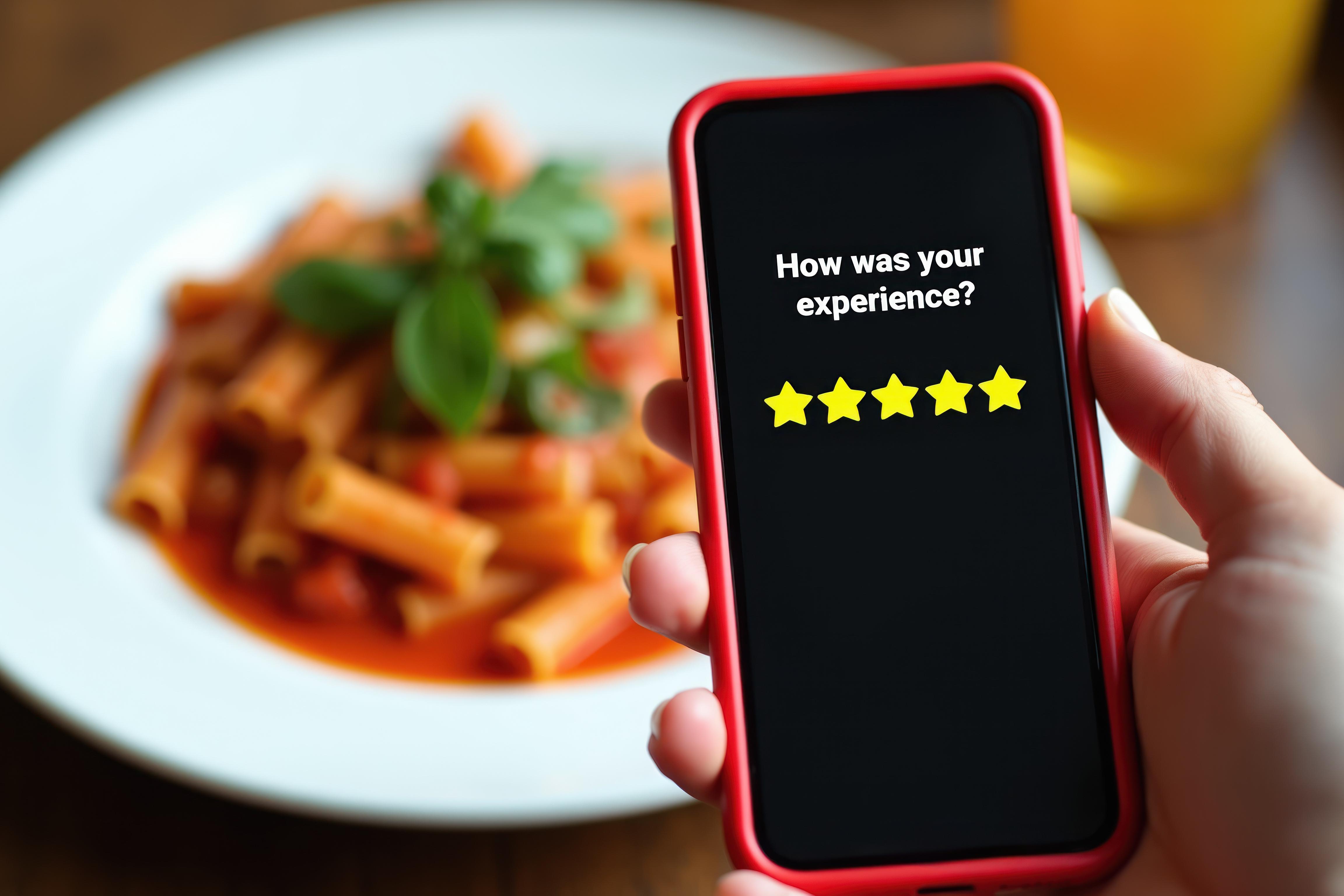 Hand holding a smartphone displaying a five-star review rating in front of a plate of pasta, illustrating an online restaurant review.