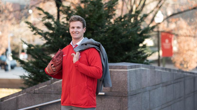 Luke Carell standing outside with red pullover, gray suit pants, red baseball glove on hand and gray suit coat draped over shoulder
