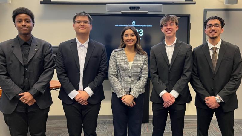 group of five students in business suits in front of screen in classroom