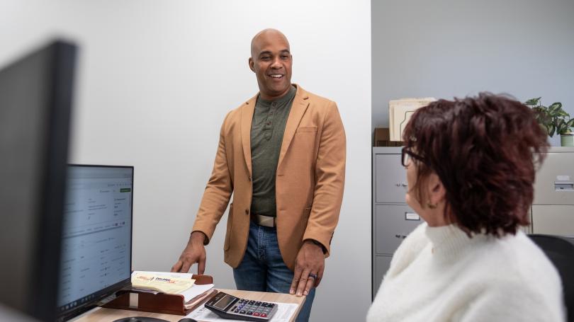 Levi Duncan and a colleague talk in an office as one stands beside a desk with a computer, papers and a calculator while the other sits facing him.