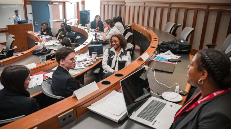 Students seated in a tiered classroom participating in a group discussion with laptops, papers, and notebooks on the desks.
