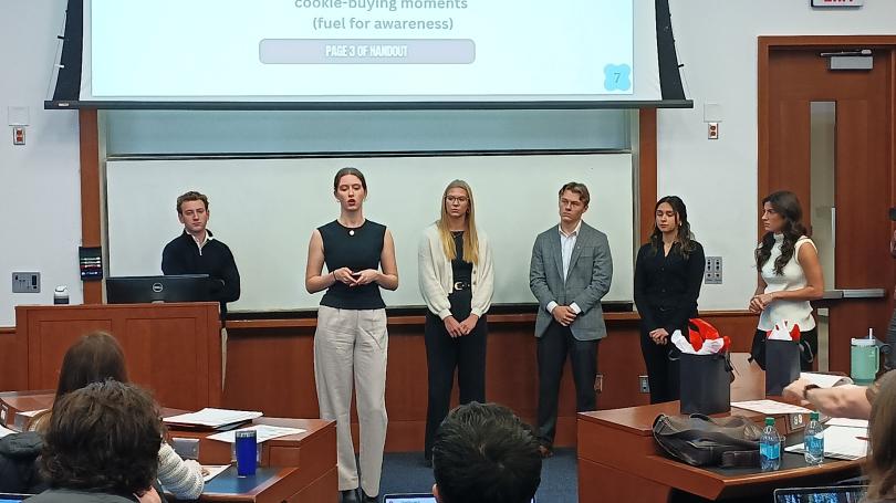 Group of students in front of classroom with presentation screen pulled down.