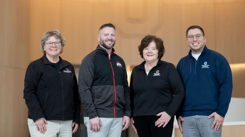 MBOE alumni from left to right, Lori Abshire, Calen Bowshier, Devanie Gossett and Mike Turnbull pose in the lobby of University Hospital in front of a wood paneled wall with a block O engraved in it.