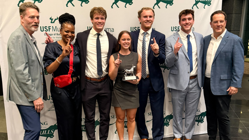Seven people holding up finger for number one in front of a sign saying "Selling with the Bulls"