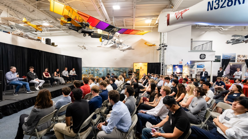 A group of alumni and students are seated on chairs in front of a panel of six people incise an aviation museum with replicas of airplanes overhead.