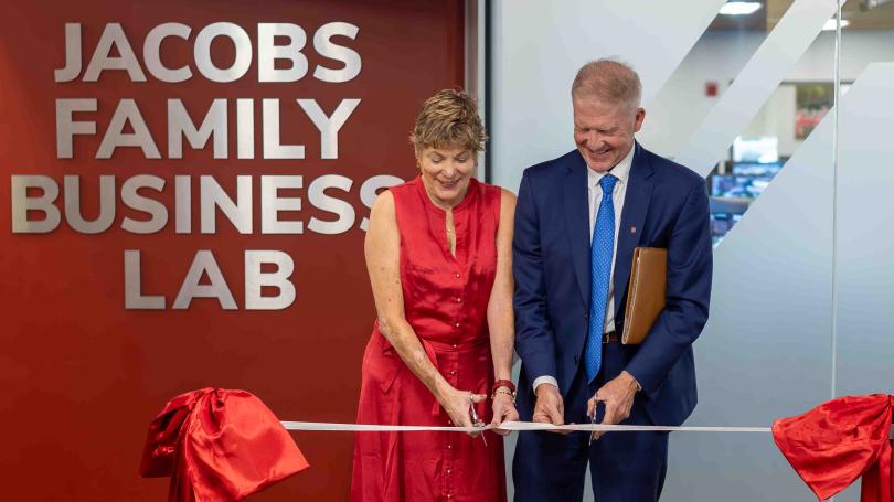 Brenda Jacobs in a red dress and Dave Jacobs in a blue suit each hold a pair of scissors and cut into a ribbon in front of the Jacobs Family Business Lab.