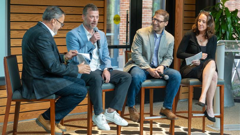 Three men and one woman sit on high stools as part of a panel discussion, with one man holding a microphone.