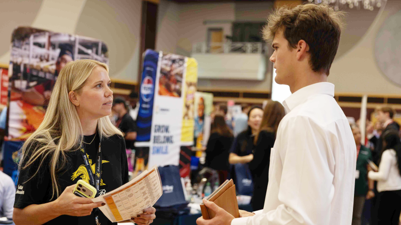Female job recruiter in black company tee shirt greets a male student in a white dress shirt and holding a folder.