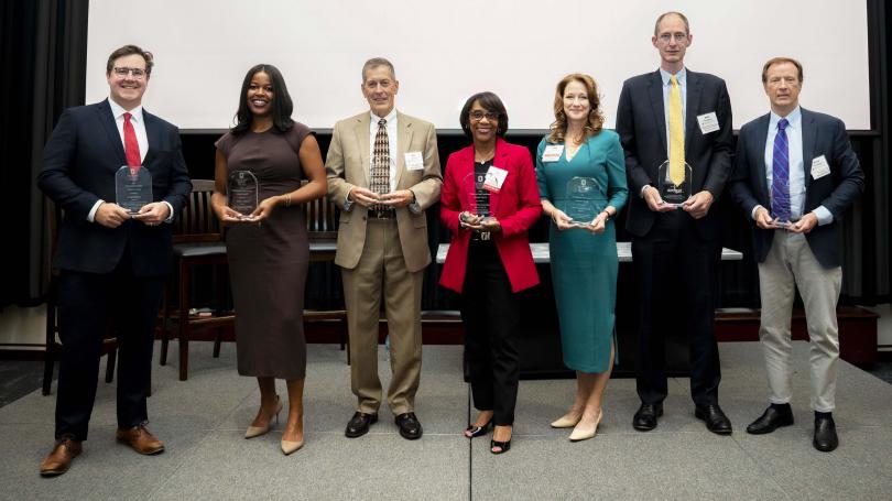 Seven men and women in business dress stand in a line holding glass awards.