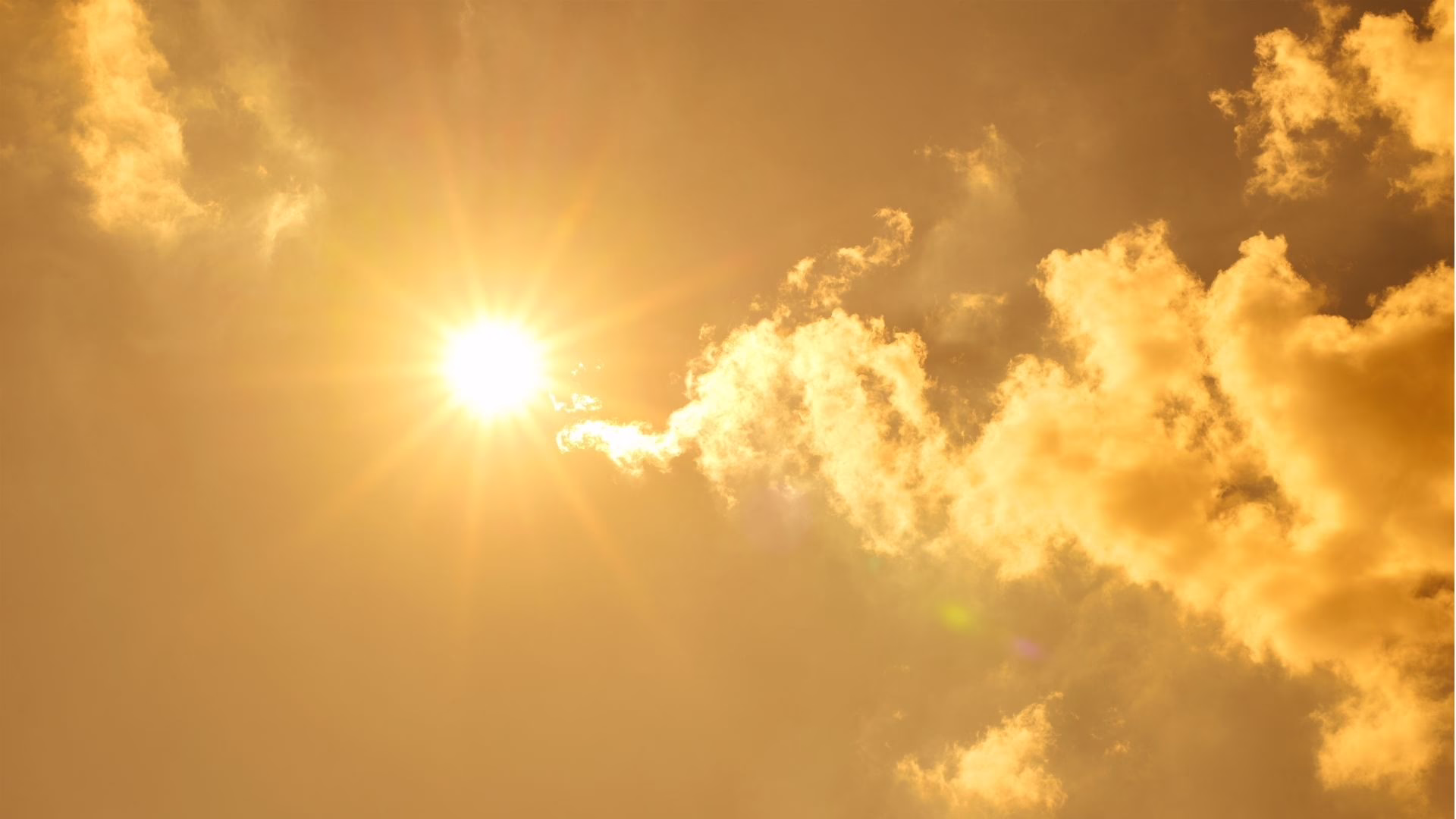 The Heat is On 7.13.26 Bright sun in a golden sky with scattered fluffy clouds