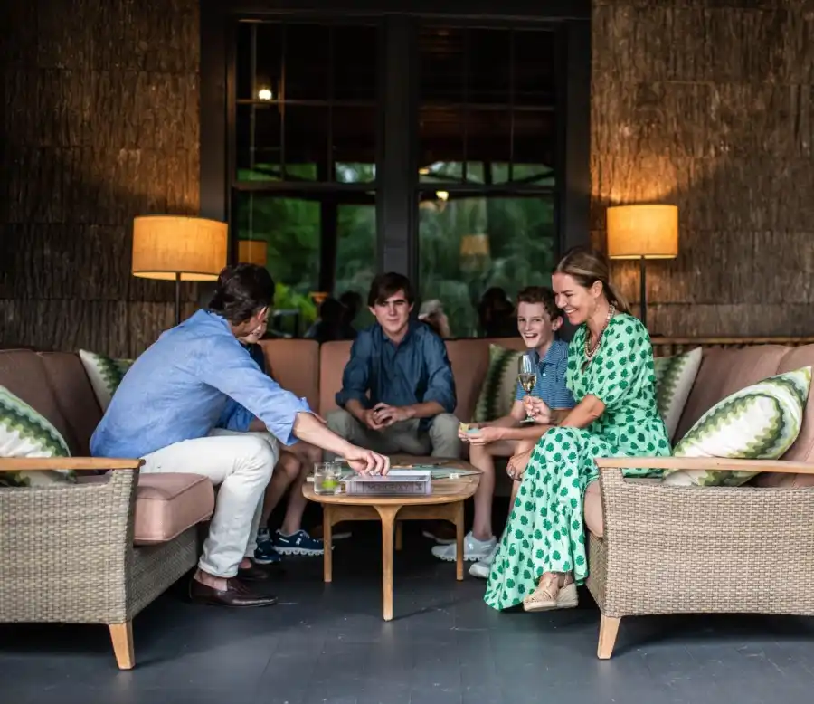 A mother, father, and their three children enjoy a lively board game in an inviting outdoor living space. Set against a backdrop of exterior windows framed by stylish, warm-toned light fixtures, the cozy scene captures the warmth of family togetherness at High Hampton Resort.