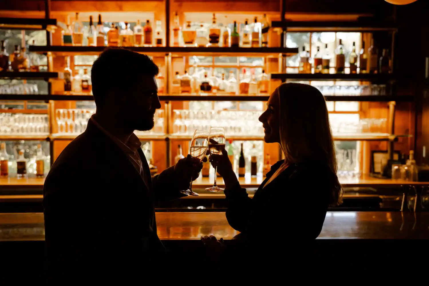 A silhouetted photo of a couple clinking champagne flutes in a toast, set against the warm glow of a softly lit bar.