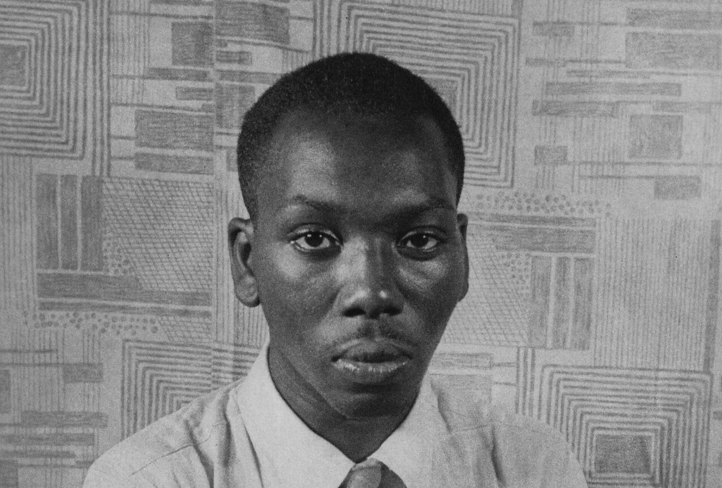 Jacob Lawrence, smiling and seated in his studio, surrounded by paints and a partially completed canvas full of bold shapes.