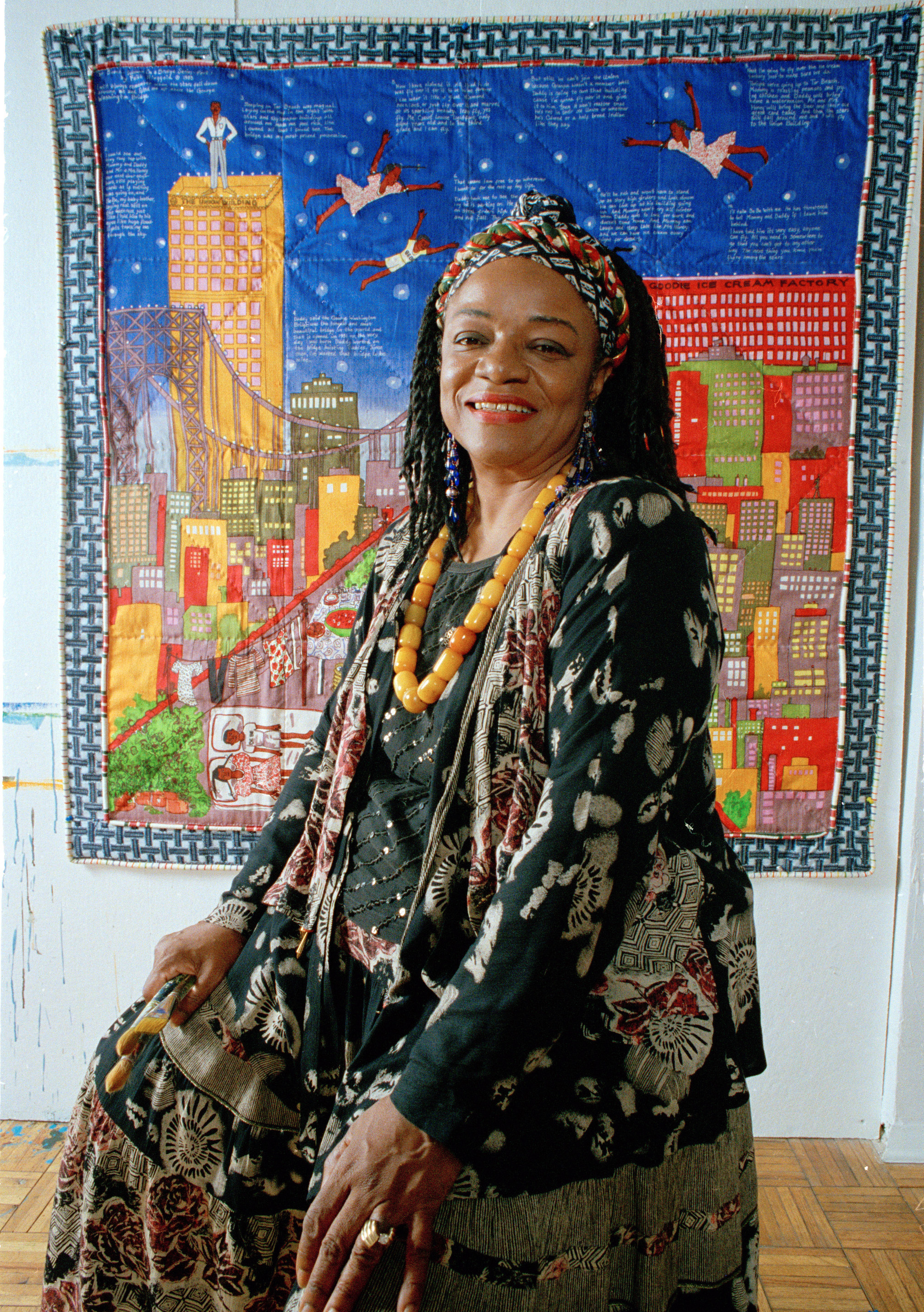 Faith Ringgold, wearing a patterned headwrap, standing beside one of her story quilts that boldly fuses art and activism.