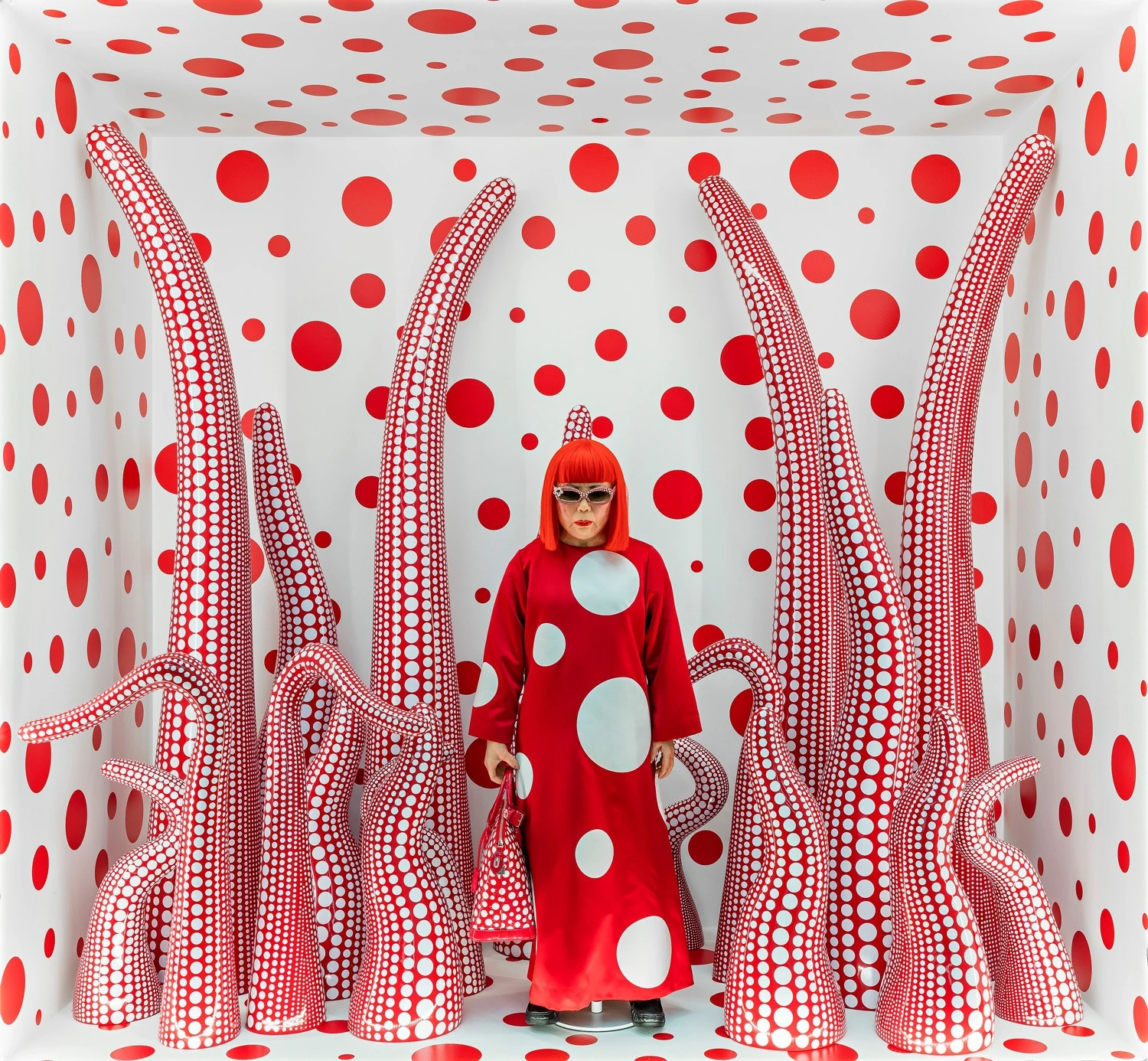 Yayoi Kusama, dressed in a polka-dotted outfit, seated among her immersive dot-covered artworks.