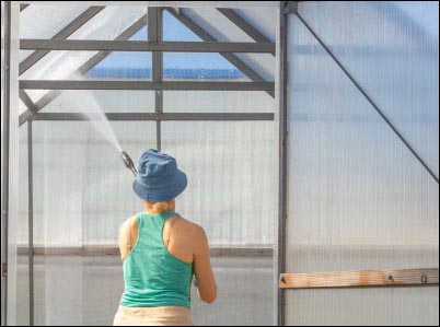 A woman using a high-pressure washer to clean the inside of a greenhouse. The image shows maintenance work in a farming environment.