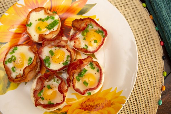 A plate of bacon and egg cups garnished with chopped green onions. The dish is served on a floral-patterned plate, with a woven mat and a bit of green fabric visible in the background.