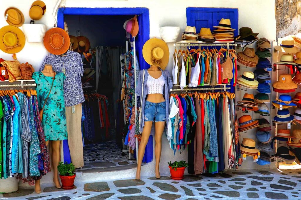 A roadside boutique store with colorful clothes on a rack and straw hats on the mannequin, by the door, and the windows.