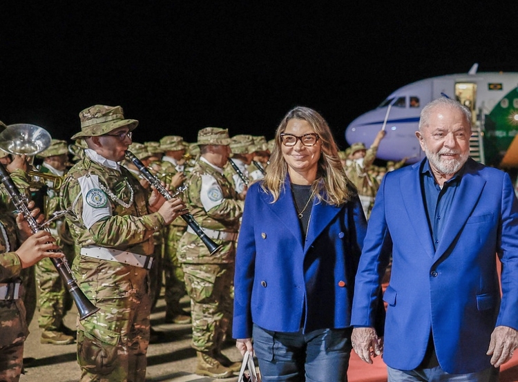 Janja e Lula de ternos azuis ouvindo a banda do exército com um avião atrás