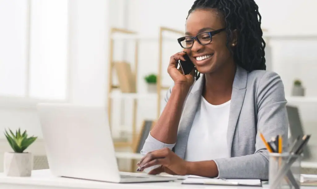 Get in Touch with Mayshine - Young woman talking on cell phone while on the computer at a desk.