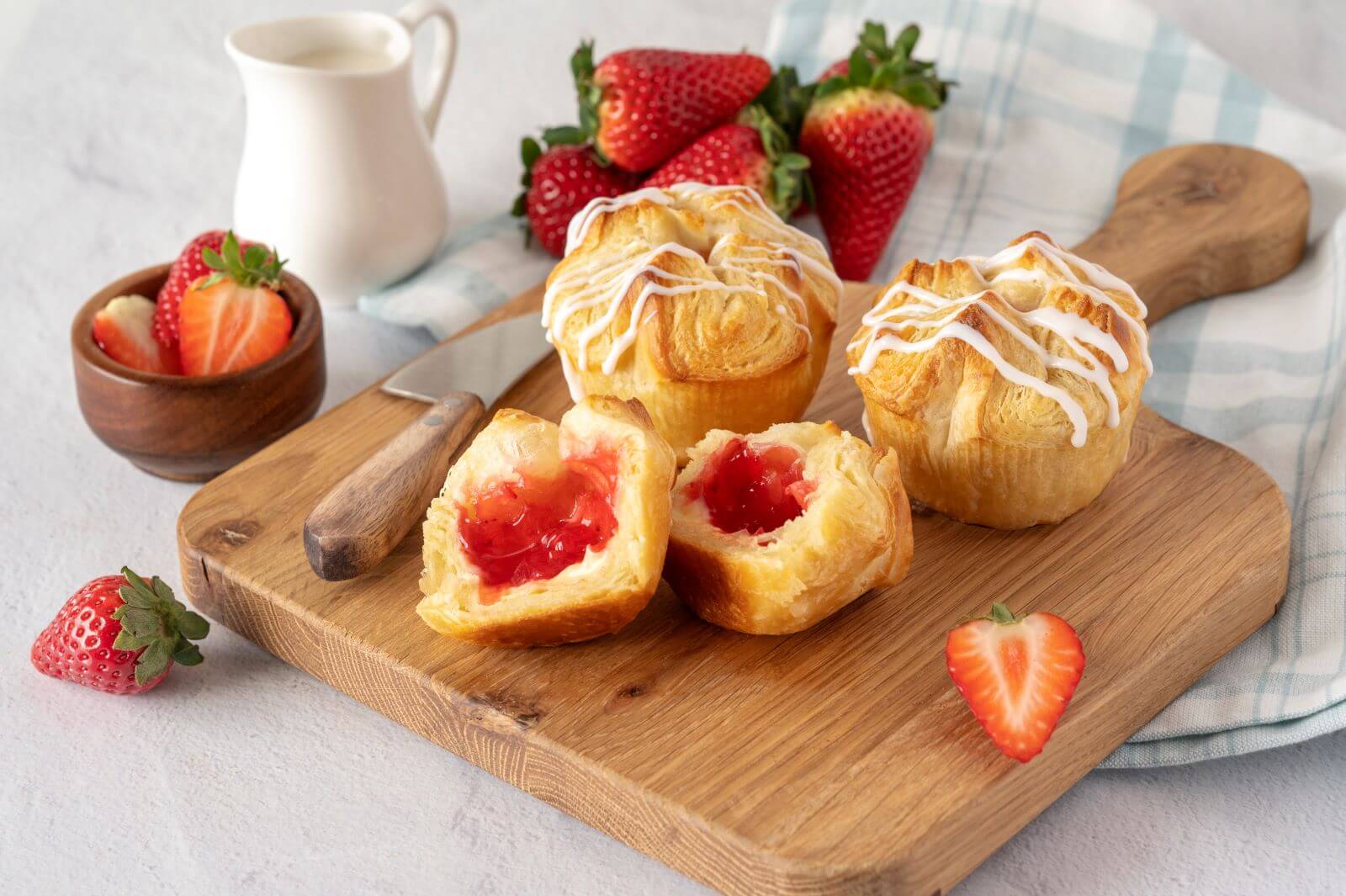 Three Strawberry & Cream Croissant Crowns on a wood cutting board. One of the crowns is cut in half. Around the crowns are slices of strawberries, a bowl of strawberries, and a creamer.