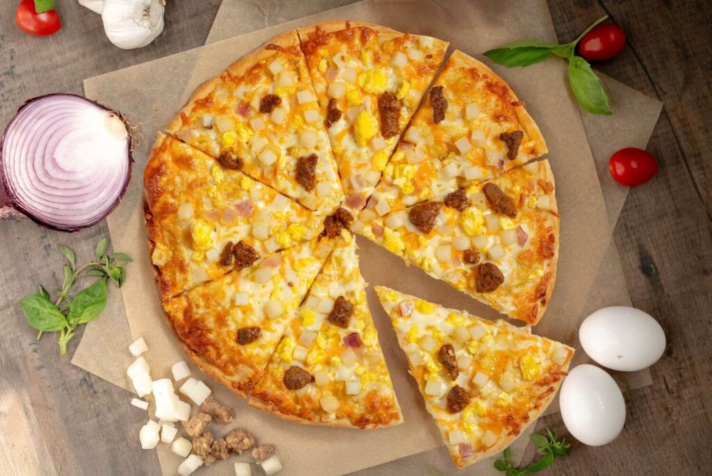 Top down photo of a baked Breakfast pizza. The pizza is on brown parchment paper with ingredients scattered all around.