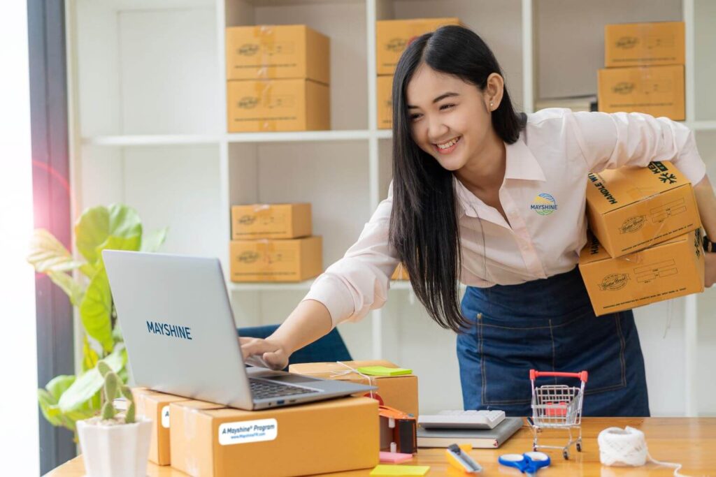 Young woman, holding two boxes and typing on a computer. More boxes on shelves behind her. Boxes are all branded with Butter Braid Pastry and Mayshine information.