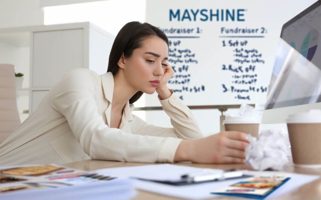 A woman leaning on her hand and holding a coffee cup while sitting at a table. Laying on the table are various fundraising marketing materials. She's tired and overwhelmed.