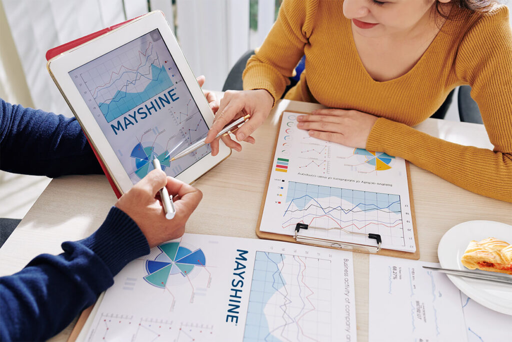 Two people looking over charts and graphs related to their fundraising event. One chart, shown on a tablet, has the Mayshine® logo. There's also a plate with a pastry slice on it.