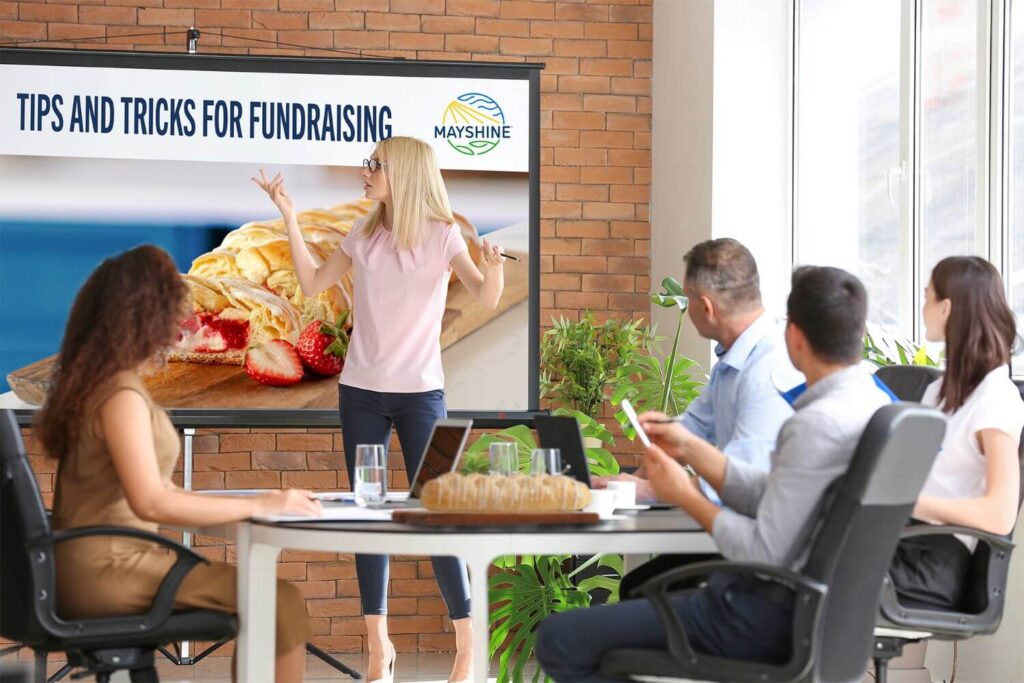 A group leader giving a presentation titled "Tips and Tricks for Fundraising" to a group of adults in a conference room. There is a Butter Braid Pastry on the table.
