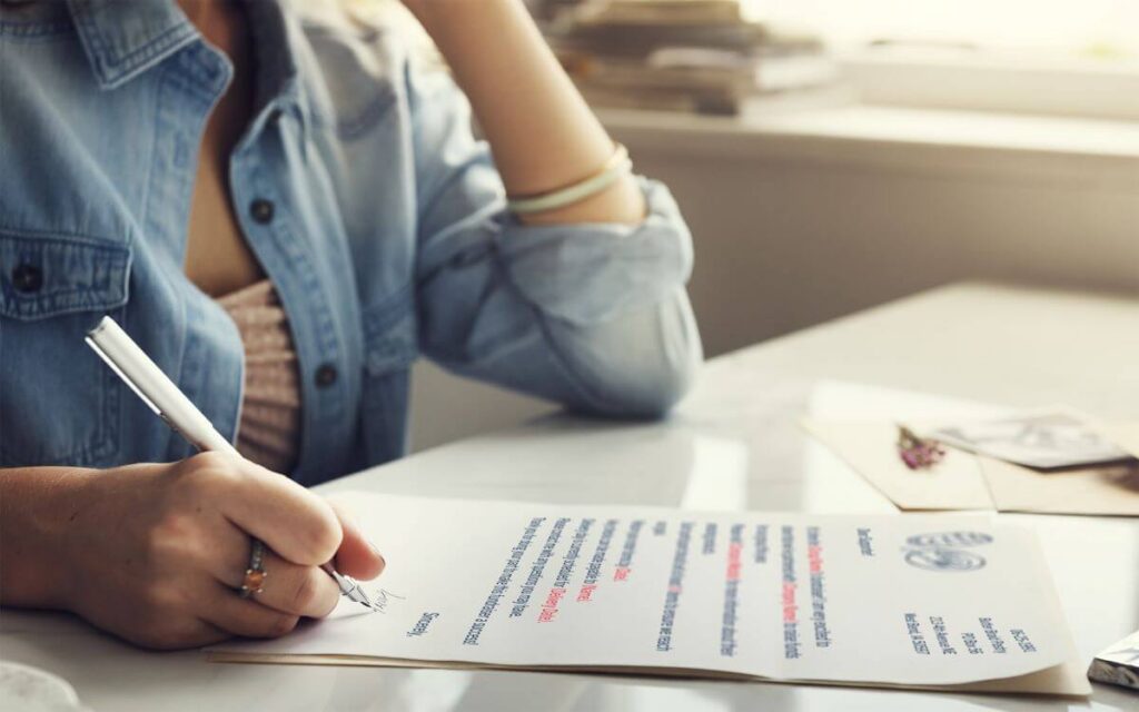 Focus is on a piece of paper on a desk. That paper is a fundraising communication piece: a parent letter. A woman is sitting at the desk, signing her name to the bottom of the letter.
