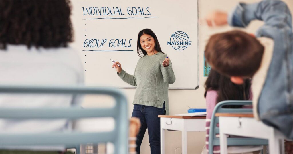 Teacher in front of a classroom writing on a whiteboard. On the board it says "Individual Goals" and "Group Goals".