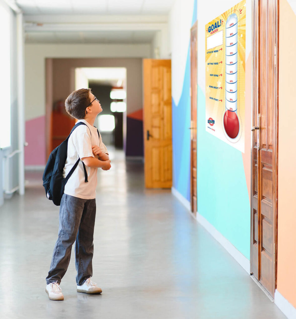 Older elementary school-aged kid looking at a fundraising goal thermometer tracker on the wall in a hallway at his school.
