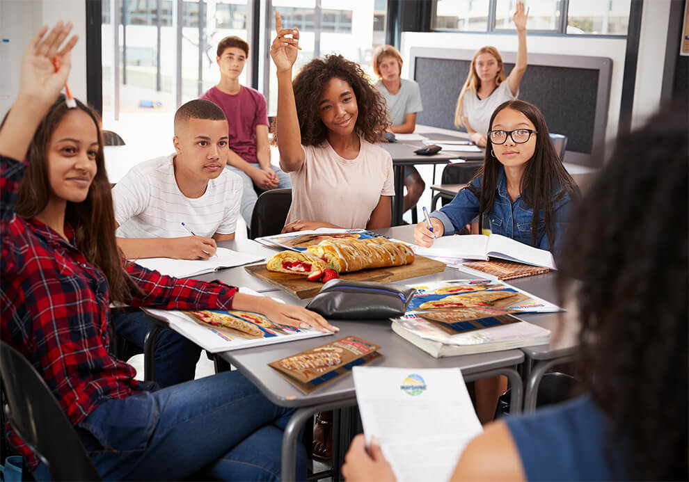 A group of high school students in a classroom looking over fundraising materials and asking their group leader questions.