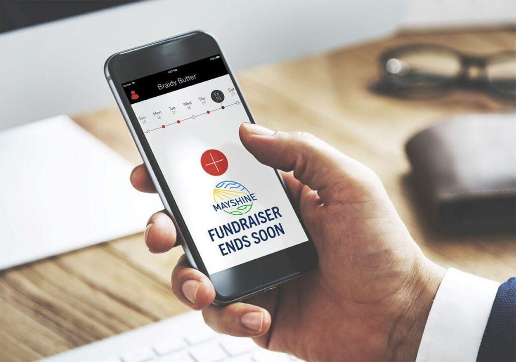 Person holding a phone with a reminder showing on their calendar. The reminder has the Mayshine logo and "Fundraiser Ends Soon" message.
