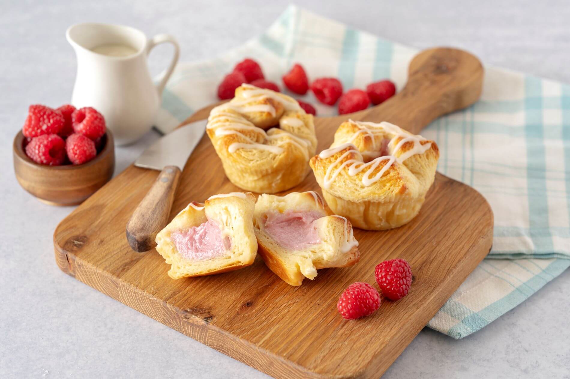 Three Raspberry Cream Cheese Croissant Crowns on a wood cutting board. One of the crowns is cut in half. Around the crowns are raspberries, a bowl of raspberries, and a creamer.