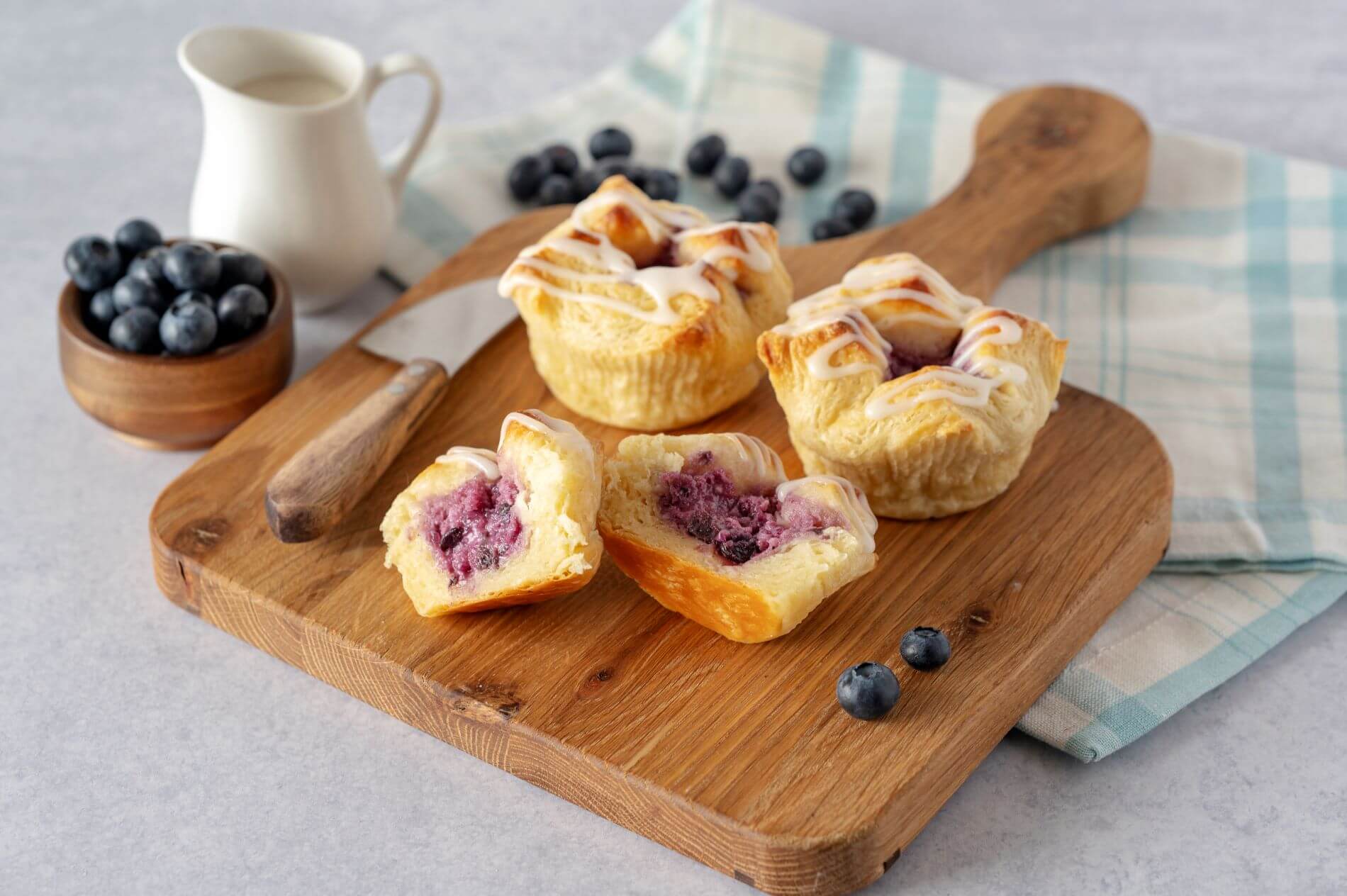 Three Blueberry Cream Cheese Croissant Crowns on a wood cutting board. One of the crowns is cut in half. Around the crowns are blueberries, a bowl of blueberries, and a creamer.