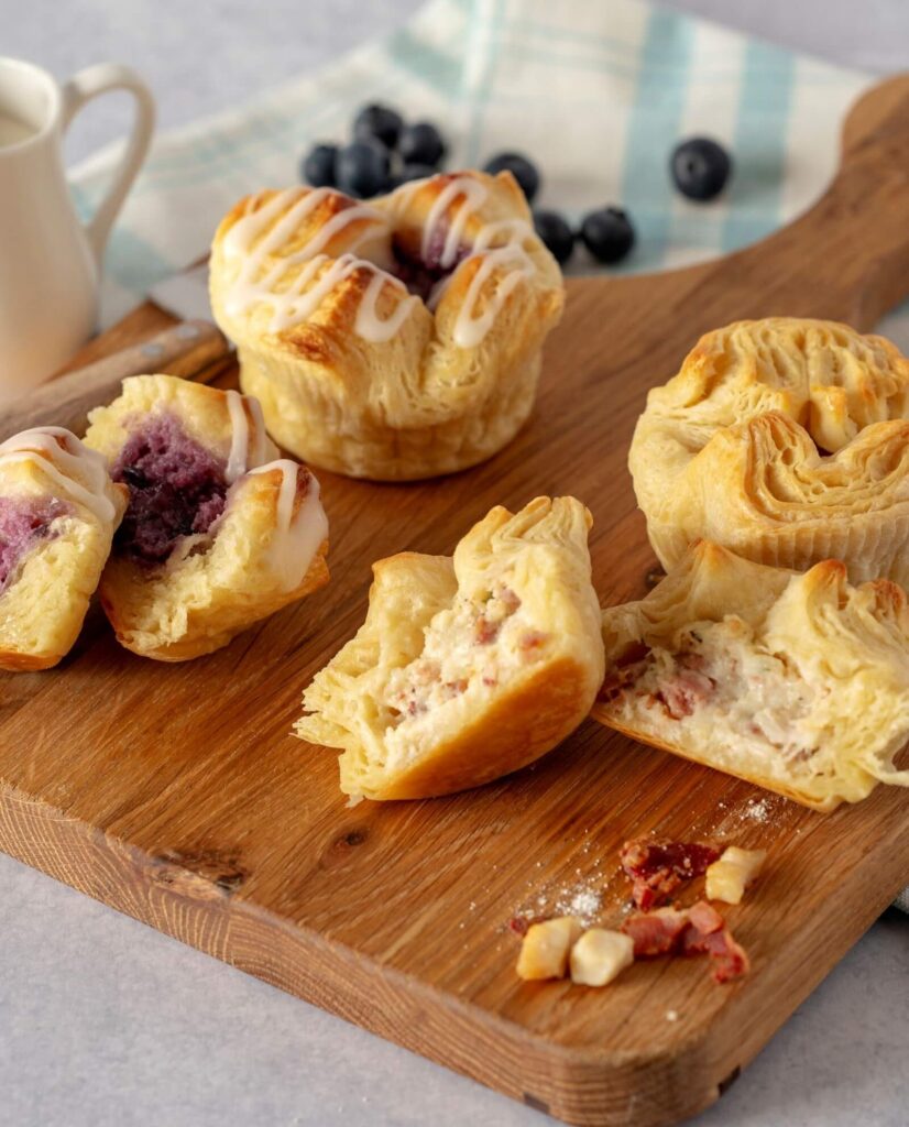 Four Croissant Crowns on a wooden cutting board. 2 are Blueberry Cream Cheese and 2 are Chicken Bacon Ranch. Several are cut in half showing the filling.