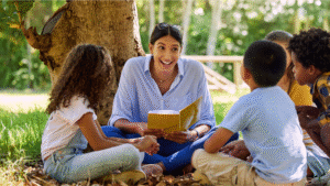 A teacher reading a book to a group of 4 students while sitting under a tree outodors