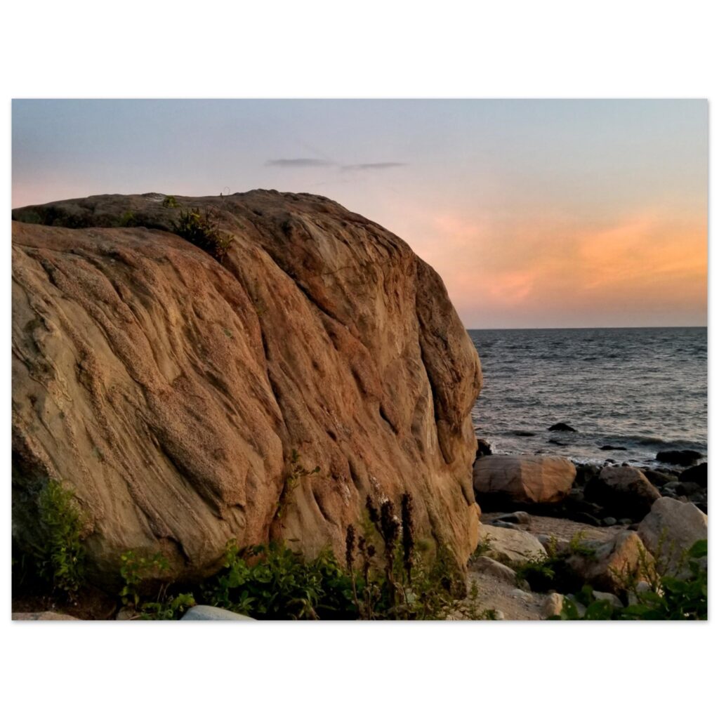 A large rock formation overlooking a calm ocean at sunset. The sky is painted in soft orange and pink hues, creating a serene and peaceful atmosphere.