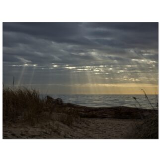 Coastal sunset with rays filtering through clouds over a calm sea. Foreground features sand dunes and grasses, conveying a serene, contemplative mood.